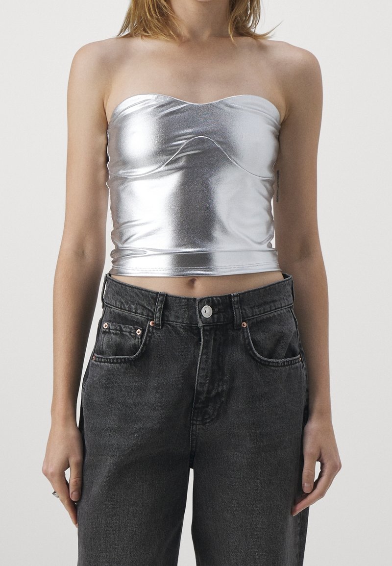 Metallisk silverfärgad strapless topp med en figurnära design, som har en böjd halslinje och en slät, glänsande textur, kombinerad med högmidjade mörka denimjeans.