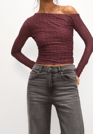 Kvinde iført en langærmet, skulderfri tekstureret bordeaux top kombineret med grå højttaljede jeans foran en ensfarvet baggrund.
