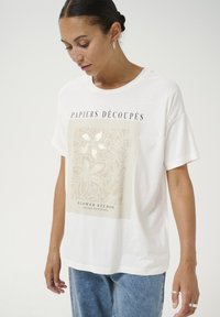 White cotton t-shirt featuring a large light beige graphic with floral patterns and text "PAPIERS DÉCOUPÉS" and "FLOWER STUDIO" in black.