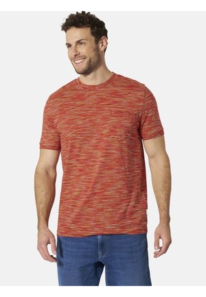 Lächelnder Mann, der ein rot-orange gestreiftes kurzärmeliges T-Shirt mit Brusttasche und blaue Jeans trägt, vor einem einfarbigen Hintergrund steht.
