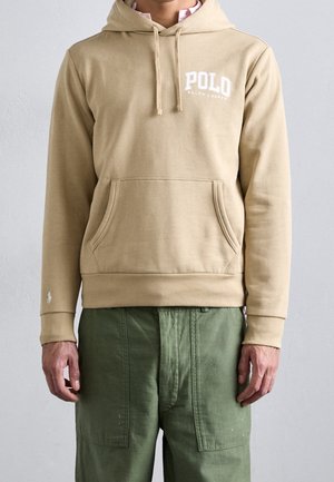 Mann trägt eine beige Polo Ralph Lauren Kapuzenjacke mit Fronttasche und olivgrüne Hose, steht vor einem schlichten hellen Hintergrund.