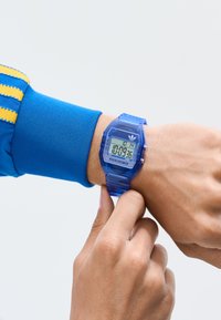Person som bär en blå digital armbandsur som visar tiden 10:09:36, justerar klockan på handleden med ett finger.