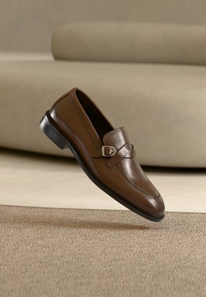 Brun skinn-loafer med detalj i sølvspenne, plassert i luften over et beige teppe og foran en beige sofa.