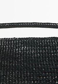 Bracelet en cuir tressé noir et surface texturée assortie posés sur un fond blanc, montrant un motif tressé détaillé.