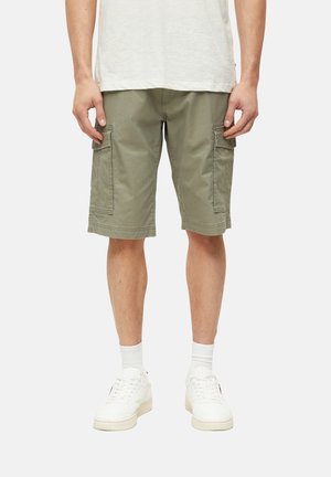 Mężczyzna ubrany w oliwkowo-zielone krótkie spodnie cargo, białe skarpetki typu crew, białe trampki oraz jasny t-shirt, z rękami spoczywającymi wzdłuż boków.