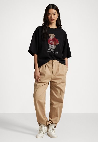 Črna oversize majica s pisanom grafiko medveda v rdečem puloverju, v kombinaciji z bež visokim pasom in belimi tenisicami.