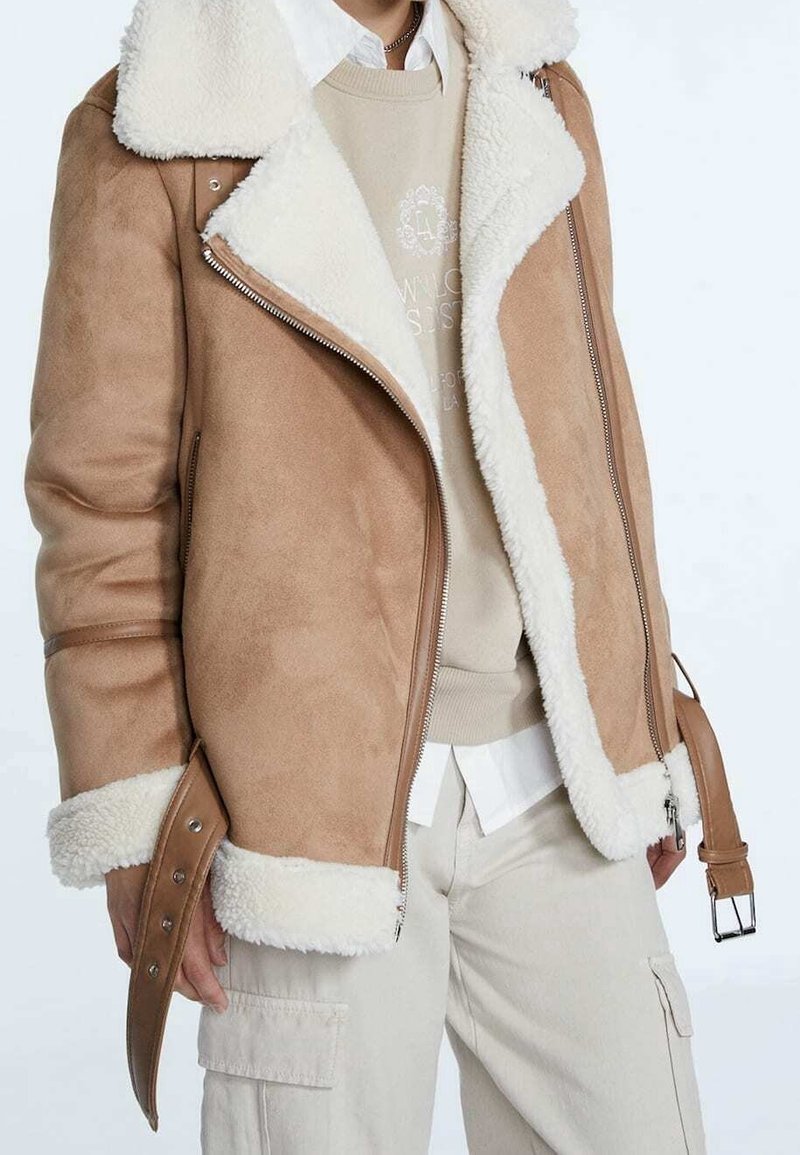 Man in een tan schapenvachtjack met brede revers, beige sweatshirt, witte shirt en lichtgekleurde cargobroek, staande tegen een effen achtergrond.