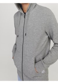 Sudadera gris con cremallera hecha de un tejido suave, con capucha, cremallera negra y bolsillos laterales. Presenta una pequeña etiqueta de logo en el borde inferior.