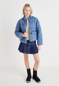 Denim puffer jacket with a high collar, light blue color, stitched detailing, paired with a dark denim skirt featuring red embroidered accents. Black ankle boots.