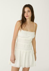 Weißes Kleid mit Spaghettiträgern, das ein gestuftes Design und strukturiertes Material aufweist, mit Rüschendetails an der Oberseite und dem Saum.