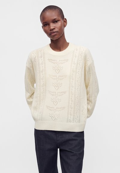 Personne portant un pull en maille crème avec un motif de feuille texturé, associé à un pantalon large foncé, debout devant un fond uni.