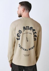 Beige langærmet skjorte med sort tekst på ryggen, herunder "LOS ANGELES" og "REVIVAL NIGHT." Blødt, bomuldsstof, afslappet pasform.