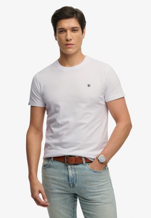 Joven con camiseta blanca lisa, vaqueros azul claro, cinturón de cuero marrón y reloj de pulsera con correa marrón, de pie frente a fondo blanco.