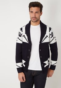 Cárdigan de punto en azul marino y blanco, con cuello chal, cierre de botones y patrones geométricos en las mangas y el cuerpo. Textura suave.