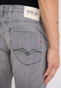 Grå denimjeans med en texturerad yta. Har ett svart sydd mönster på bakfickan och en vit märkesetikett vid midjan.