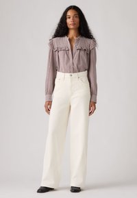 Γυναίκα με μακριά σγουρά μαλλιά φοράει ένα καρό πουκάμισο με βολάν, μπα tucked μέσα σε λευκό παντελόνι με φαρδιά μπατζάκια και ψηλή μέση, και μαύρα παπούτσια, στέκεται ουδέτερα.