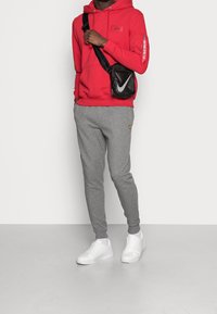 Červená mikina s kapucí a předním logem, v kombinaci se šedými zúženými tepláky s malým logem a bílými teniskami. Součástí je černá crossbody taška.
