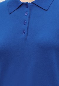 Gros plan d'un polo en maille bleue avec un col pointu et quatre boutons bleus assortis sur la patte de boutonnage avant.