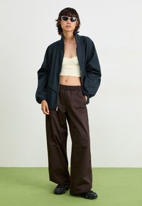 Sort letvægts bomberjakke, cremet cropped top, brune afslappede bukser med elastisk talje og sorte chunky sko. Grøn gulvbaggrund.