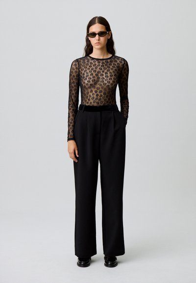 Femme portant un haut noir transparent à manches longues avec des fleurs, un pantalon large taille haute noir, des mocassins noirs et des lunettes de soleil sombres, se tenant devant un fond uni.