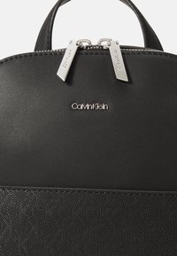Čierny kožený batoh s embosovaným geometrickým vzorom, striebornými zipsami a logom Calvin Klein. Hladký povrch, zaoblený tvar a dvojité madlá.