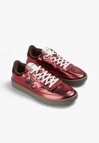 Zapatillas deportivas rojas metálicas con superficie texturizada, cordones blancos y lengüeta marrón; cuentan con un emblema de calavera y tibias cruzadas en el lateral.