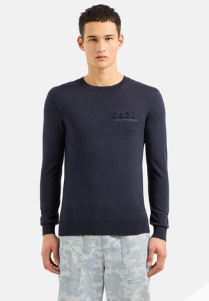 Maglione blu navy a maniche lunghe con collo rotondo, polsini a coste e un logo testurizzato sul lato sinistro del petto. Realizzato in un materiale a maglia liscia.