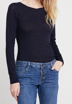 Vrouw draagt een aansluitende doorschijnende zwarte top met lange mouwen, ingestopt in een hoog getailleerde blauwe spijkerbroek met vier zichtbare knopen.