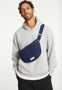 Sac bandoulière en denim bleu avec un dessus zippé, une forme arrondie et une sangle réglable, porté sur un sweat à capuche gris. L'étiquette sur le sac indique le nom de la marque.