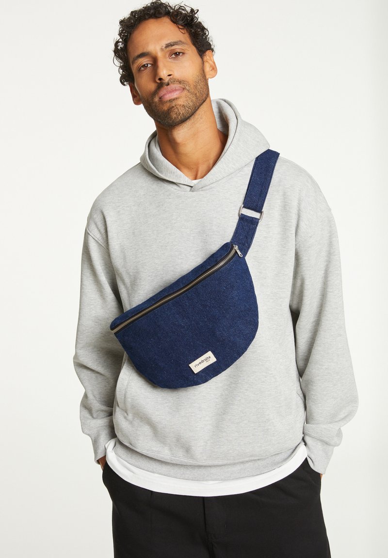 Sac bandoulière en denim bleu avec un dessus zippé, une forme arrondie et une sangle réglable, porté sur un sweat à capuche gris. L'étiquette sur le sac indique le nom de la marque.