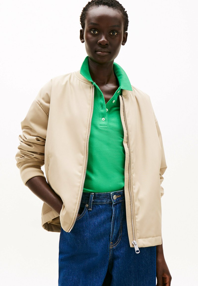 Giubbotto bomber beige con zip frontale, polsini a costine e polo verde sotto. Abbinato a jeans in denim blu. Tessuto liscio e vestibilità comoda.