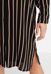 Svart skjortklänning med vertikala svarta, vita och bruna ränder; har knappar längs framsidan och sidoklyftor för detalj.