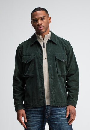 Green corduroy jacket with two chest pockets, black buttons, and a zippered beige sweater underneath, paired with blue jeans.