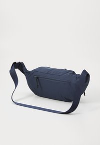 Mørkeblå pose-lignende veske laget av myk tekstil, med justerbar stropp og en glidelåslomme på forsiden for sikker oppbevaring.