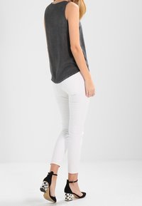 Donna che indossa un top senza maniche grigio scuro, pantaloni skinny bianchi e tacchi con cinturino alla caviglia neri con tacchi a blocco a pois bianchi, in piedi su un pavimento bianco.