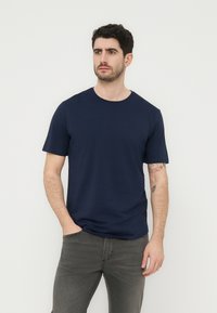 Άνδρας με σκούρα μαλλιά που φοράει μπλε σκούρο κοντομάνικο μπλουζάκι και γκρι τζιν, στέκεται με το ένα χέρι στη τσέπη μπροστά σε μονόχρωμο φόντο.