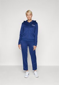 Tumši zila sporta tērpa komplekts no mīkstas auduma, kurā ietilpst džemperis ar rāvējslēdzēju un pieguļošas bikses, abiem ar baltajiem "adidas" logo uz krūšu un augšstilba.