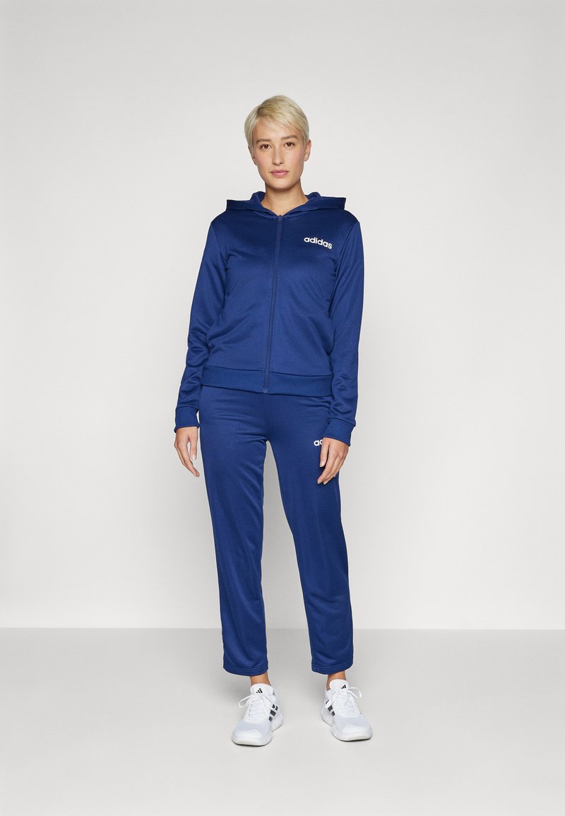 Tumši zila sporta tērpa komplekts no mīkstas auduma, kurā ietilpst džemperis ar rāvējslēdzēju un pieguļošas bikses, abiem ar baltajiem "adidas" logo uz krūšu un augšstilba.