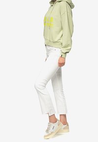 Persoon met een lichtgroene hoodie met gele tekst, witte cropped jeans en wit-beige sneakers, staand tegen een witte achtergrond.