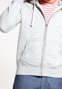 Hombre vestido con una sudadera con cremallera gris sobre una camiseta de rayas rojas y blancas y pantalones azules, ajustando la capucha con una mano.