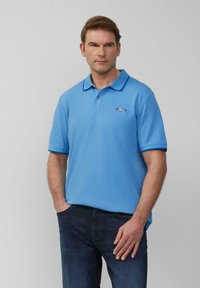 Man in een lichtblauwe polo met Ford-logo en een donkere spijkerbroek, staande met zijn handen in zijn zakken tegen een effen grijze achtergrond.