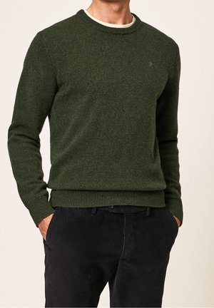 Uomo che indossa un maglione girocollo verde scuro e pantaloni neri, con le mani in tasca, mostrato dalla nuca ai fianchi su sfondo semplice.