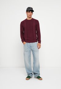 Suéter de malha em bordeaux com decote redondo, combinado com jeans de perna larga azul claro e ténis verde escuro com solas castanhas.