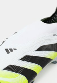 Crampons de soccer blancs avec des rayures noires, tige texturée avec un motif en grille et un accent jaune vif sur la semelle.