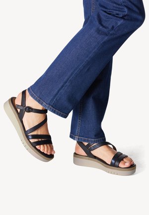 Sandales en cuir noir avec plusieurs brides, une boucle ronde et une semelle en wedge grise texturée. Portées avec un pantalon en denim bleu.