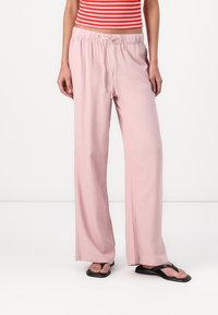 Pantalon large rose clair avec une taille élastique et un cordon de serrage. Texture lisse, design décontracté, associé à des tongs noires.