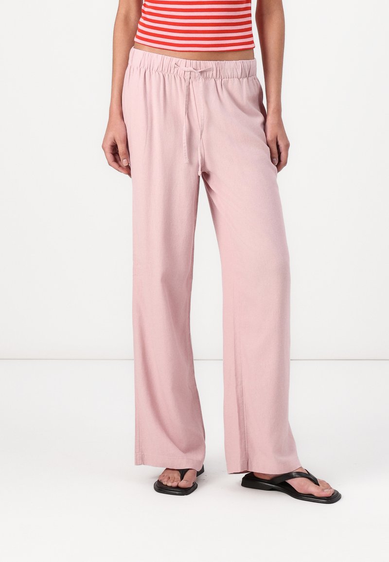 Pantalon large rose clair avec une taille élastique et un cordon de serrage. Texture lisse, design décontracté, associé à des tongs noires.