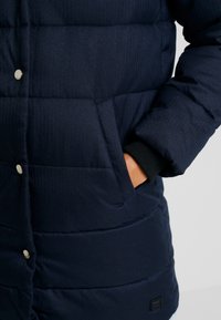 Marinblå puffjacka med quiltat mönster, utrustad med sidoficka, silverfärgade tryckknappar och ribbade ärmslut. Strukturerad tyg.