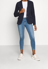 Blazer bleu marine à deux boutons, associé à un jean skinny bleu clair et des baskets blanches, créant une tenue décontractée mais structurée.