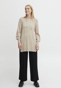 Haut à capuche beige à manches longues avec une coupe ample, un col à cordon, des poignets côtelés et un ourlet droit, associé à un pantalon large noir et des chaussures noires.